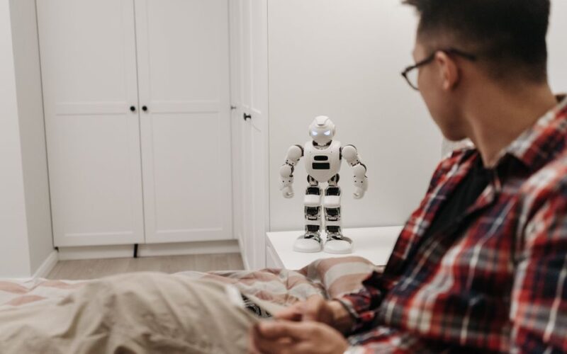 A man examines a robotic toy placed on a table in a contemporary bedroom setting.