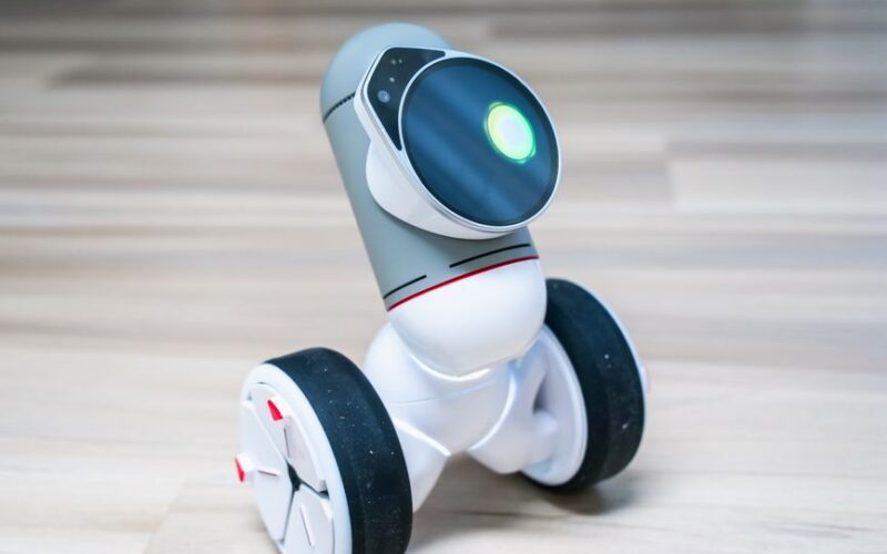 A small wheeled robot with a glowing light on a wooden floor indoors.