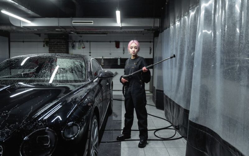 A woman using a pressure washer to clean a black sports car at an indoor car wash facility.