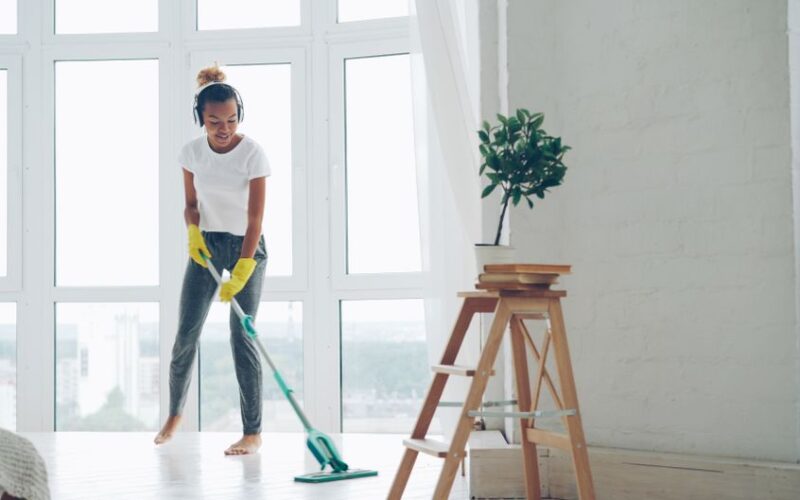 Young woman cleaning floor with a mop in a sunny room with large windows.