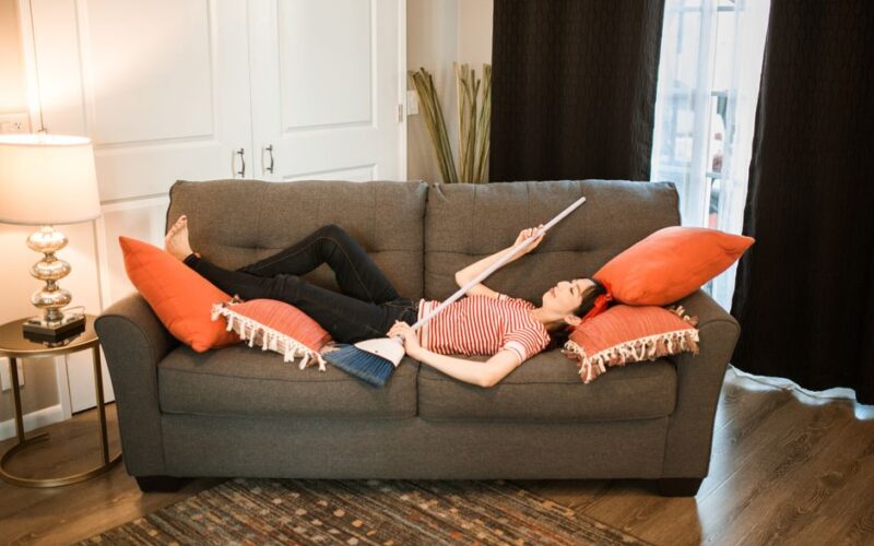 A woman in casual attire resting on a sofa indoors, holding a broom and surrounded by pillows.
