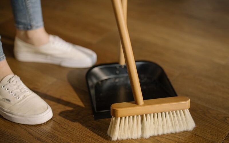 Close-up of cleaning process with broom and dustpan beside sneakers on a wooden indoor floor.