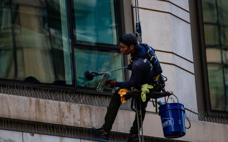 Man cleaning high-rise building windows using safety harness outdoors in daylight.