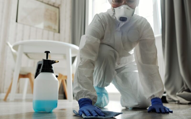 Person in protective gear cleaning a floor with disinfectant spray and cloth indoors.
