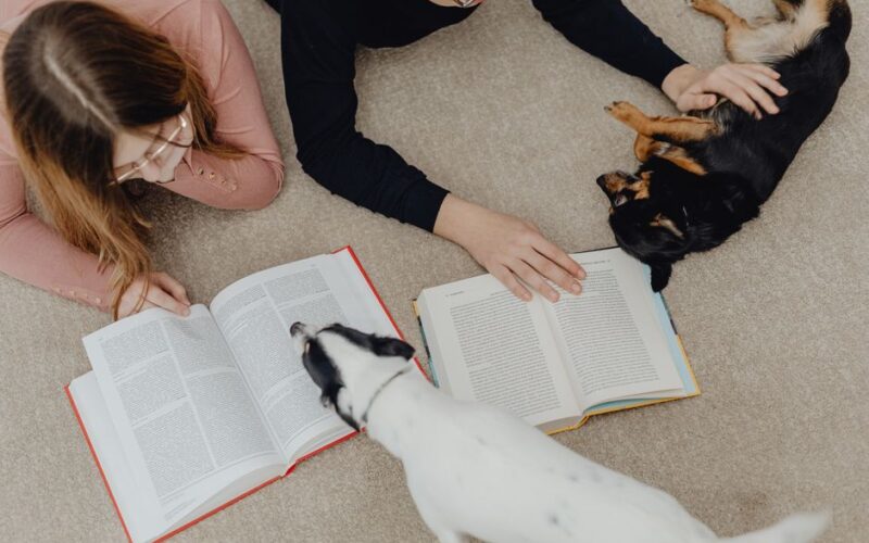 Two children reading books on the carpet while playing with cute dogs.