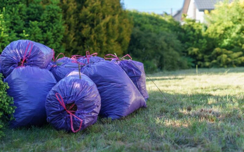 Vibrant purple trash bags stacked in a sunlit garden setting, ideal for spring clean-up visuals.