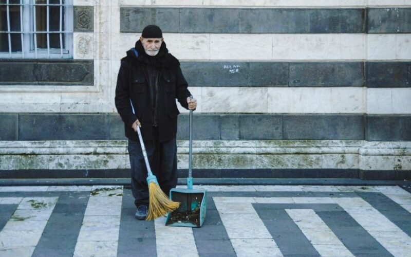 Elderly man cleaning with a broom and dustpan against a patterned marble wall.