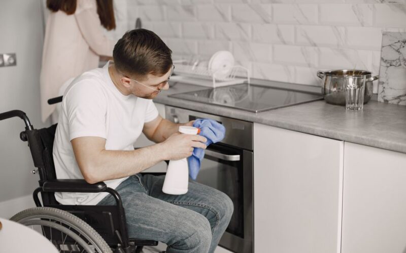 A man using a wheelchair cleans a modern kitchen surface with a spray bottle and cloth.