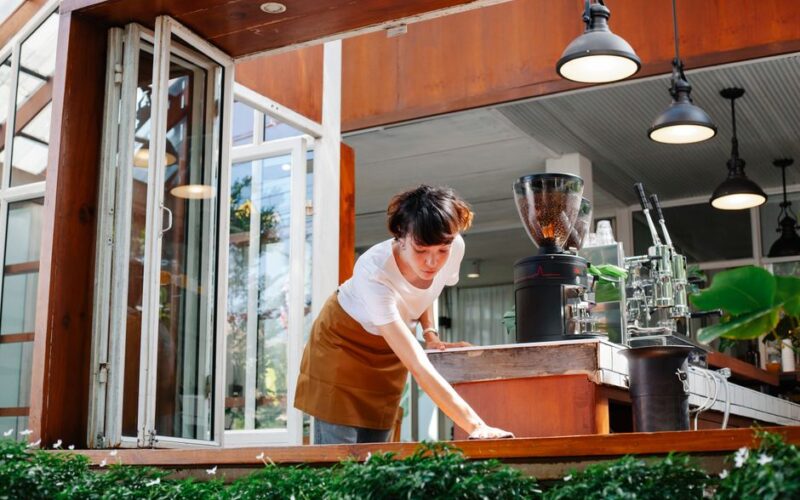 Young waitress in casual clothes and apron standing in modern cafeteria and cleaning wooden bench in daytime