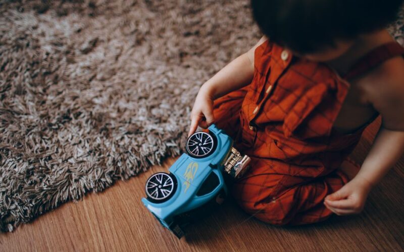 A young child in red overalls playing with a blue toy car on a carpeted wooden floor.