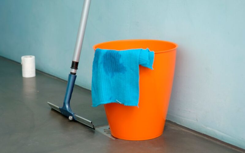 Close-up of cleaning supplies including orange bucket, mop, and blue rag indoors.