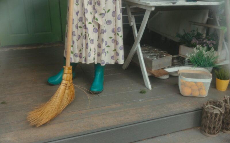 A woman in a floral dress and rubber boots sweeps a wooden porch beside a table with plants.