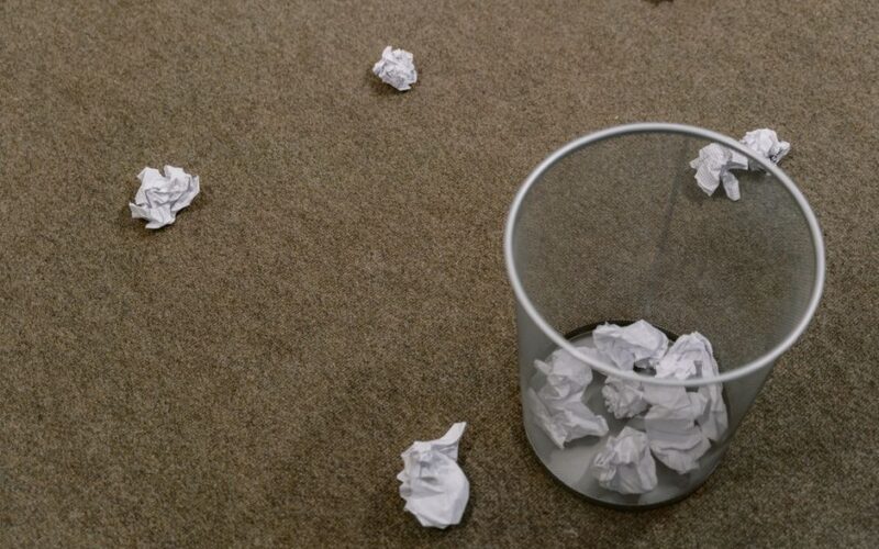 Crumpled paper scattered around a trash bin on a carpeted floor, indoors scene.