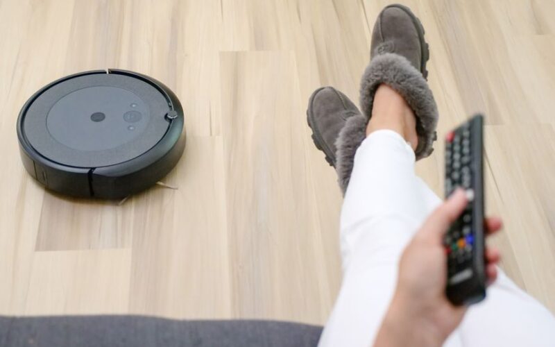 A person relaxing on a couch with a robotic vacuum cleaning the hardwood floor.