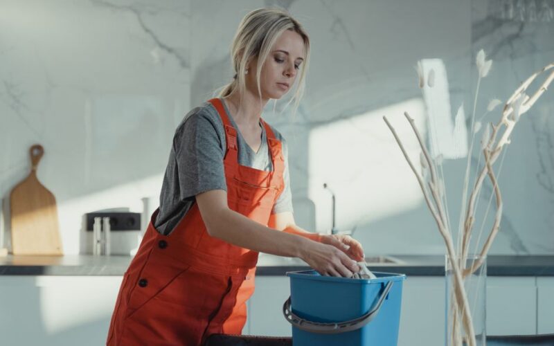 A woman in a gray shirt and red overalls cleans a kitchen counter using a blue bucket and white rag. Modern setting.