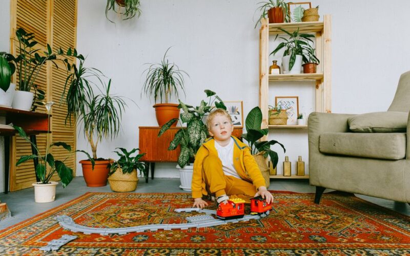 Playful child in casual wear sitting on carpet in cozy living room and playing with plastic railway and train while having fun during weekend