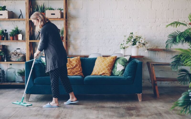 Woman with mop cleaning cozy living room with blue sofa and plants.
