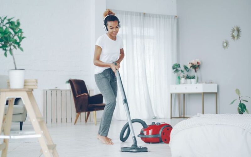 A woman in a cozy room uses a vacuum cleaner while enjoying music on headphones.