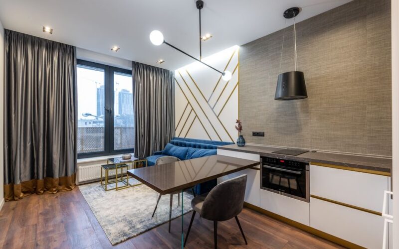 Interior of bright modern flat with table and chairs near kitchen with cabinet and counter with oven and stove near blue couch on carpet and window with curtains in daytime
