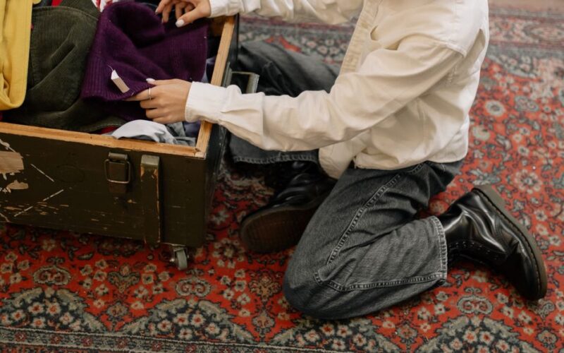 Man in white long sleeves sorting clothes from a wooden box on a patterned carpet.
