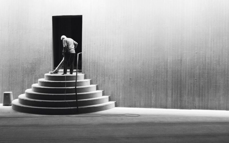 A solitary man sweeps a tiered staircase in a black and white minimalist setting.