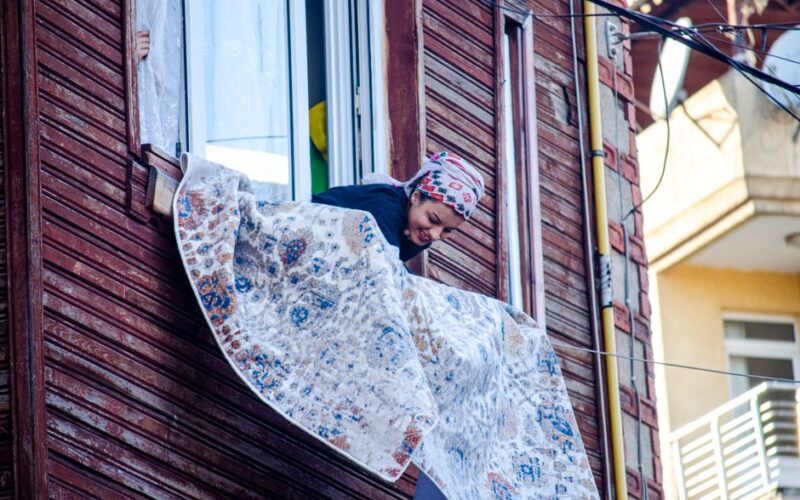 An adult woman cleaning a carpet outside a window in a vibrant city setting.