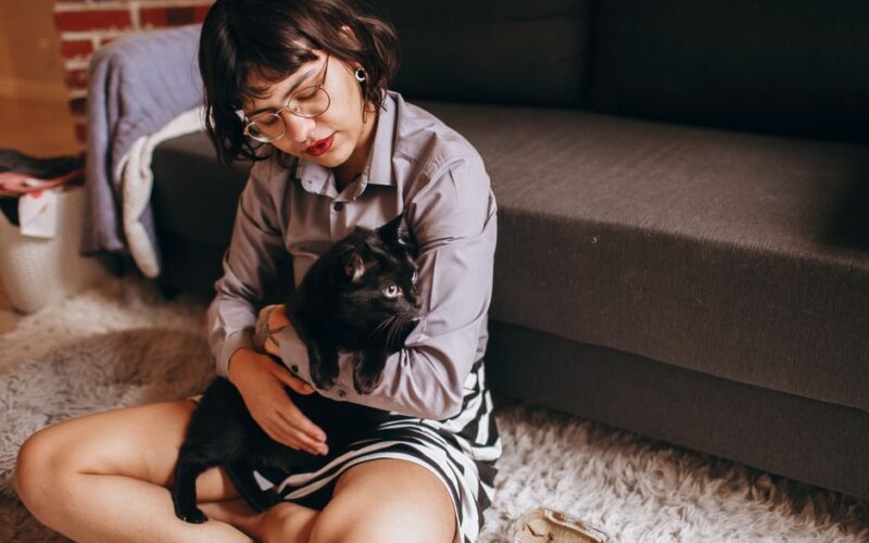 Woman holding a black cat while sitting on a cozy rug indoors.