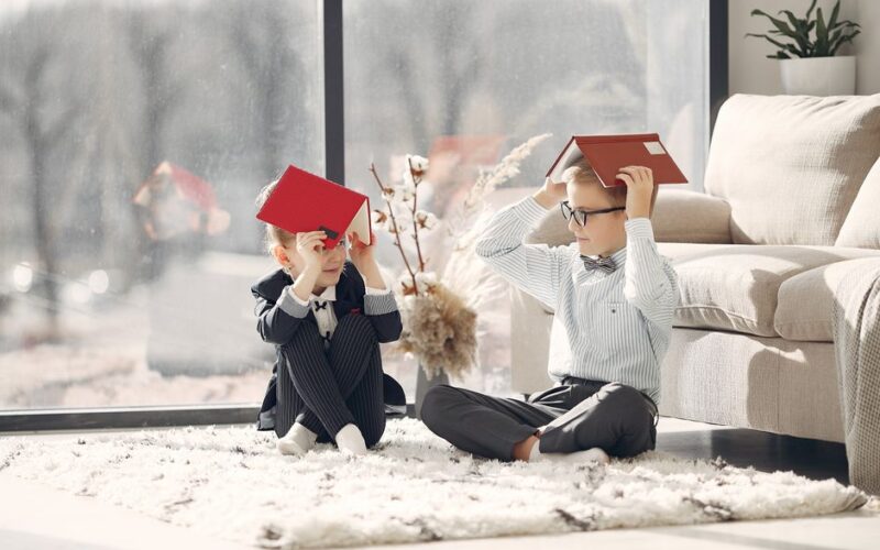 Full body little freelancers in uniform putting notebooks on head while having break in cozy living room together sitting on carpet looking at each other