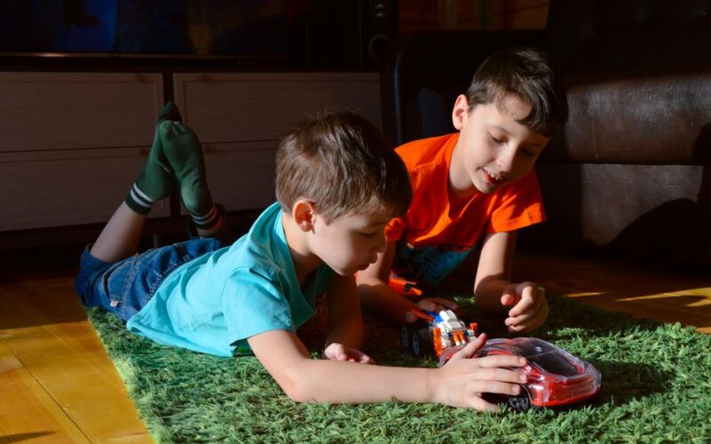 Content child with brother playing with toy auto on soft rug while resting in house room in sunlight