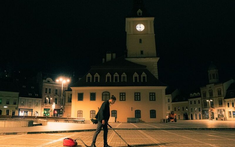 A person stands with a vacuum cleaner in Brasov's Council Square at night, amidst the historic architecture.