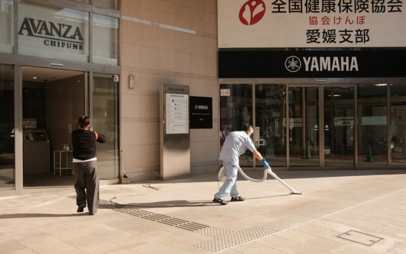 Two people clean the pavement outside a Yamaha building in Japan. Urban maintenance scene.