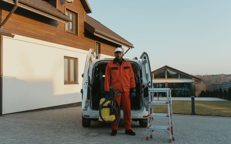 A male worker in red coveralls with a vacuum cleaner beside a van on a sunny day.