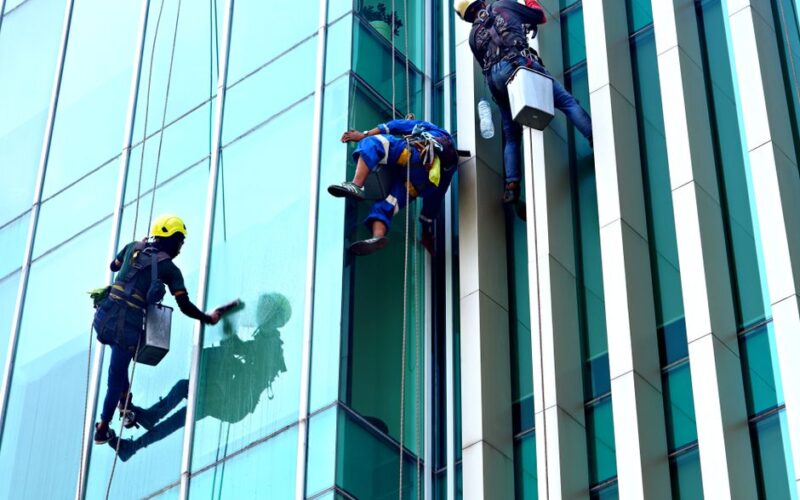 Three window cleaners suspended on ropes clean a modern glass building facade during the day.
