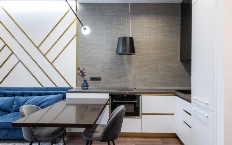 Interior of modern apartment with table with chairs near blue sofa on carpet near kitchen with counter and cabinet with stove and oven near vase with flowers