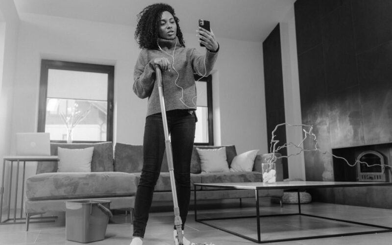 Woman multitasking by mopping the floor and video calling in a modern living room.