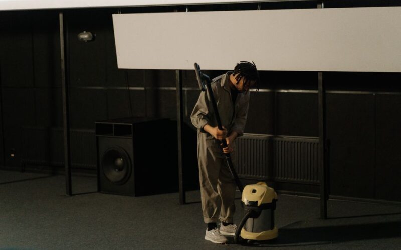 A person in uniform using a vacuum cleaner inside an auditorium with a visible speaker.