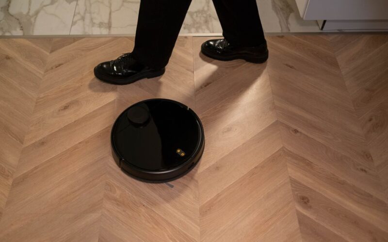A modern robot vacuum cleaner navigating wooden floors while a person in black shoes walks past.