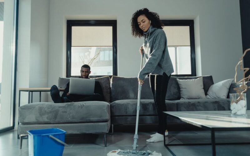 A woman mopping the floor while a man works on a laptop in a cozy modern living room.