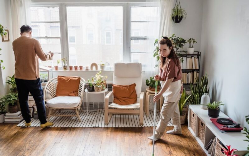 Couple cleaning a bright and cozy living room filled with plants and natural light.