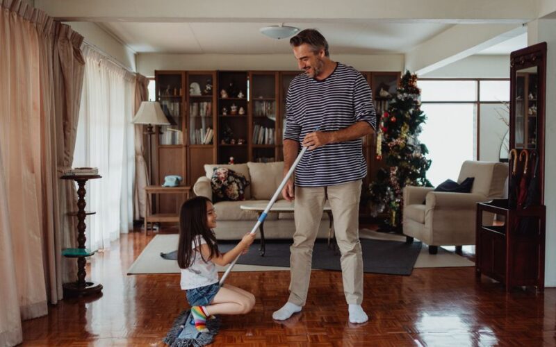Father and daughter happily cleaning and bonding at home, surrounded by a cozy living room setting.