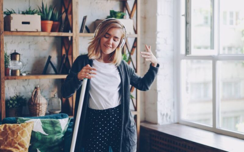 Smiling young woman with headphones dances with a broom indoors, enjoying music during cleaning.
