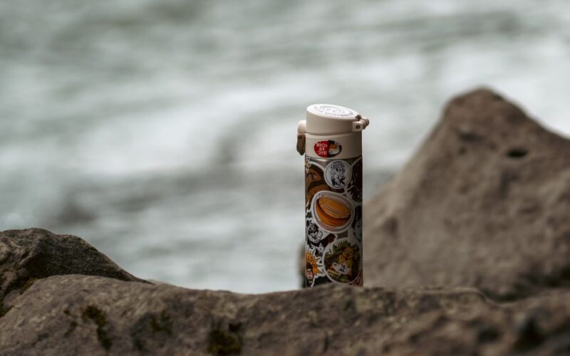 A vacuum flask decorated with stickers rests on rocks by a seashore in Russia.