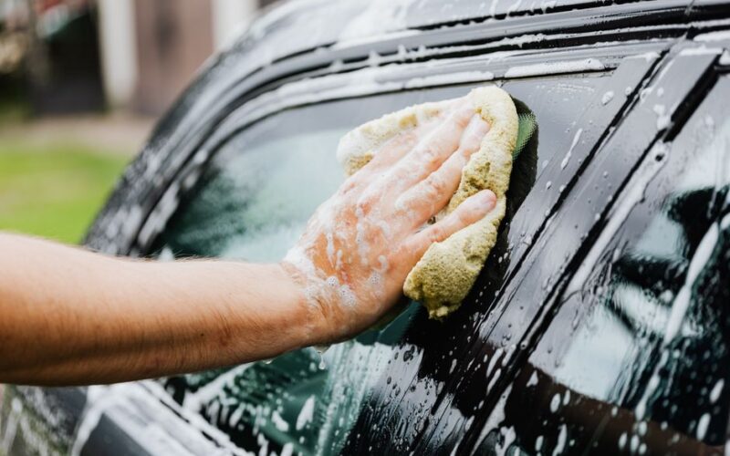 A hand scrubs a car window with a sponge, covered in soap suds, during a car wash.