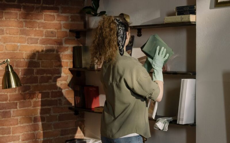 A woman dusts a bookshelf in a cozy home setting with exposed brick walls, wearing latex gloves.