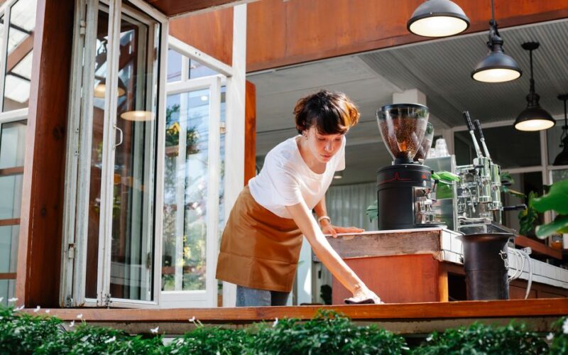 From below of focused female in apron cleaning wooden counter of modern coffee house with coffee makers and special equipment