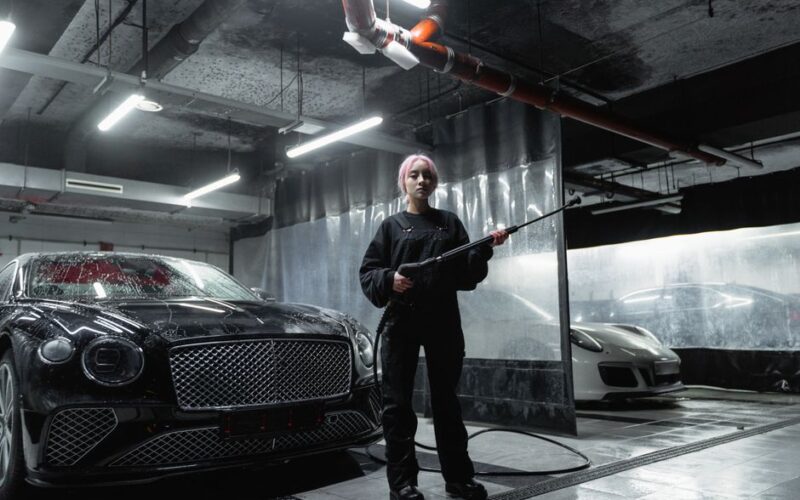 A woman in a car wash space using a power washer to clean luxury cars, captured indoors.