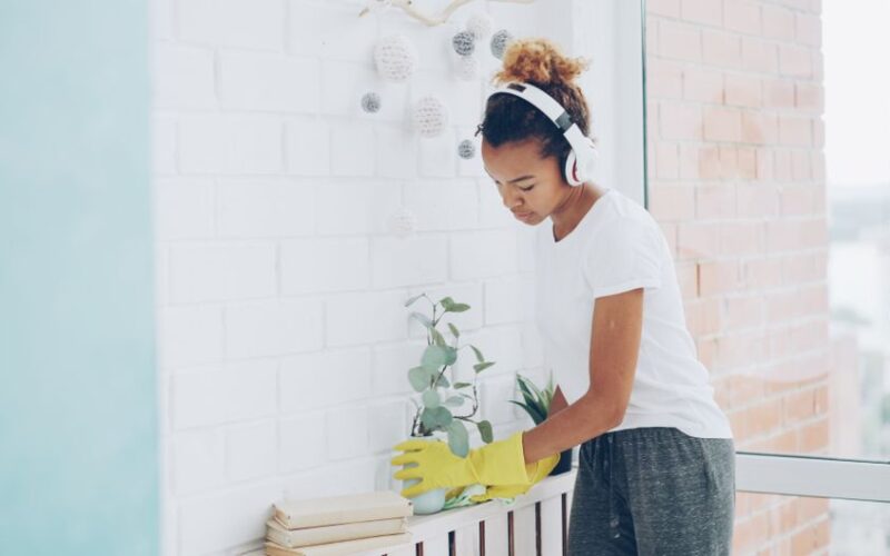 Woman wearing headphones cleaning with plant and books in bright home interior.