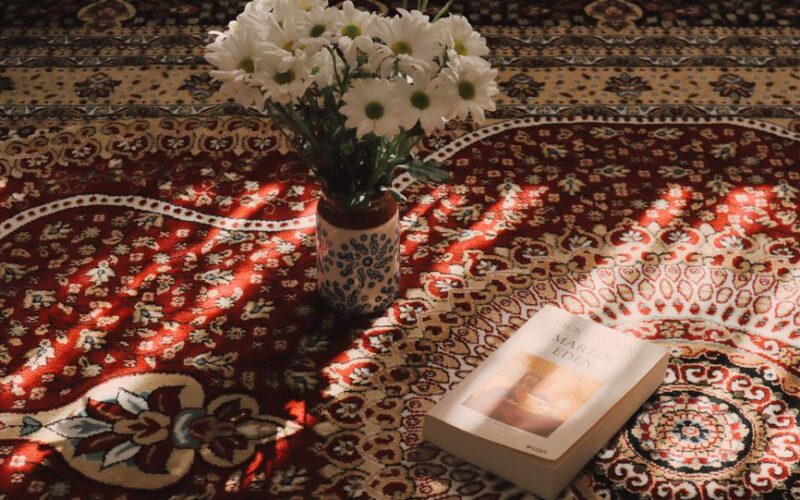 A cozy scene with white flowers in a vase and a book on a patterned carpet illuminated by sunlight.