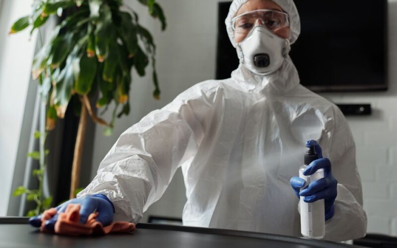 A person in protective gear cleans an indoor surface with disinfectant spray.