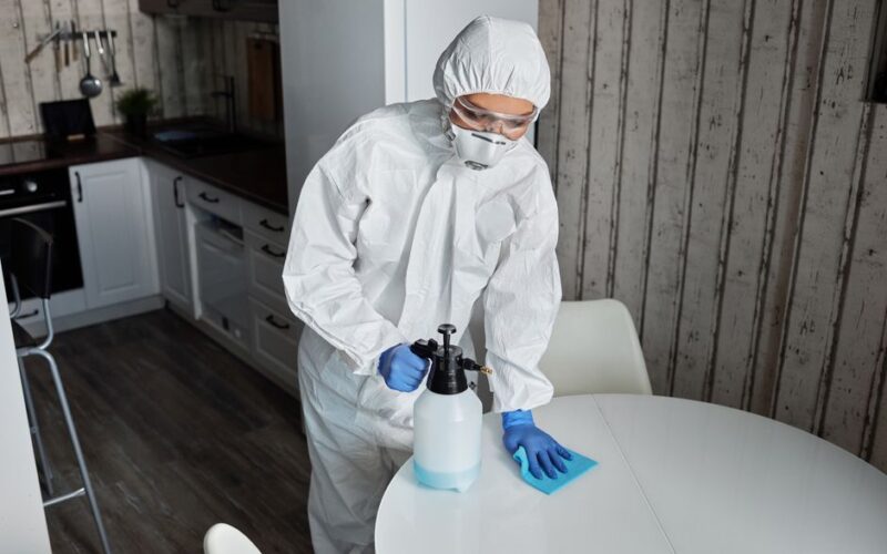 Person wearing PPE cleans a table indoors with sanitizer during pandemic.